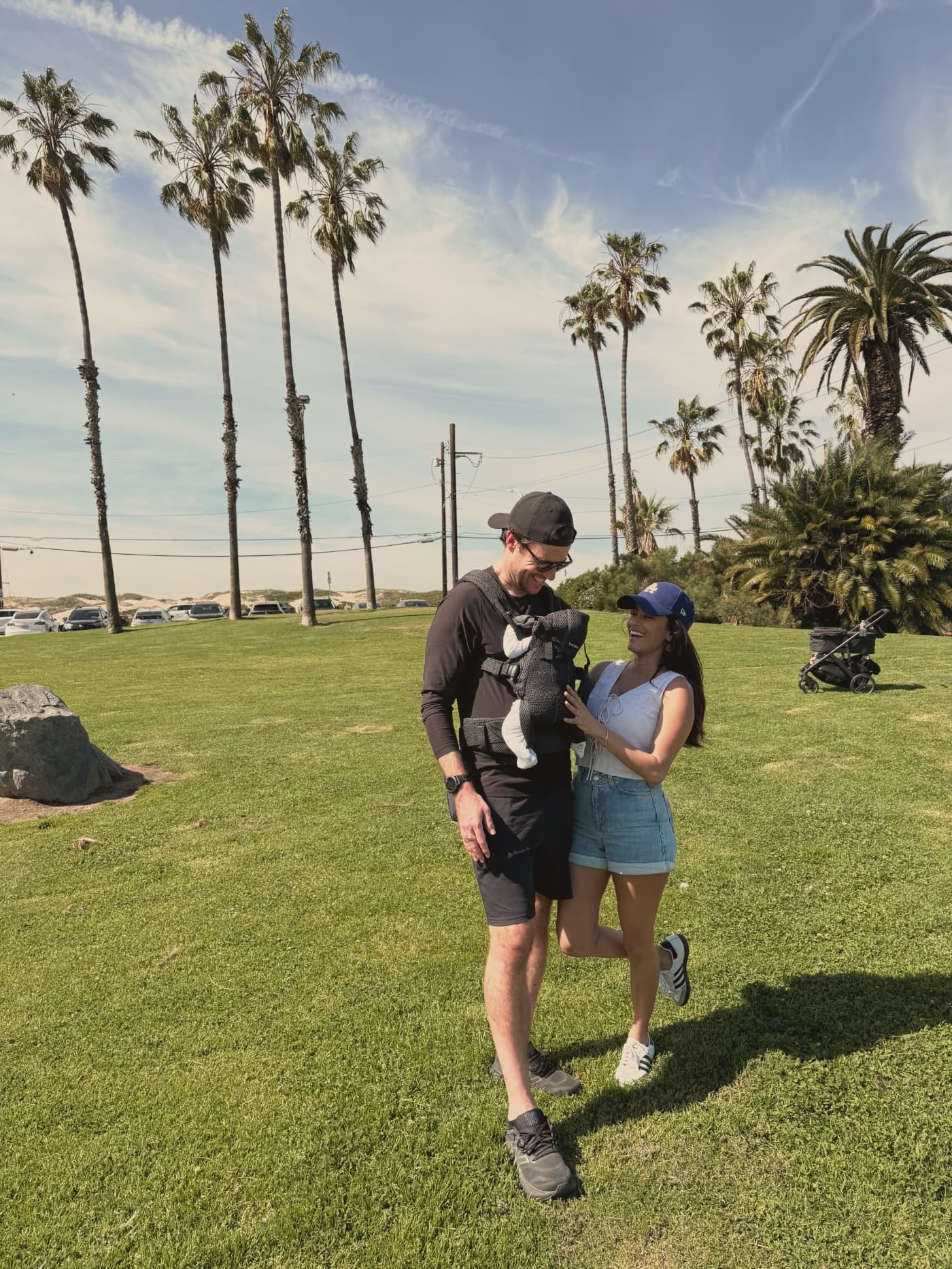 A young family outside in Los Angeles, palm trees behind them, a parent with their baby in a front carrier.